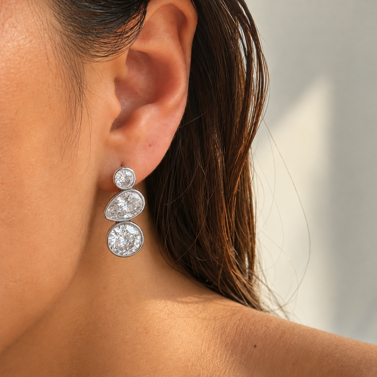 A close-up of a woman's ear wearing a silver-toned drop earring with three stacked oval clear gemstones.