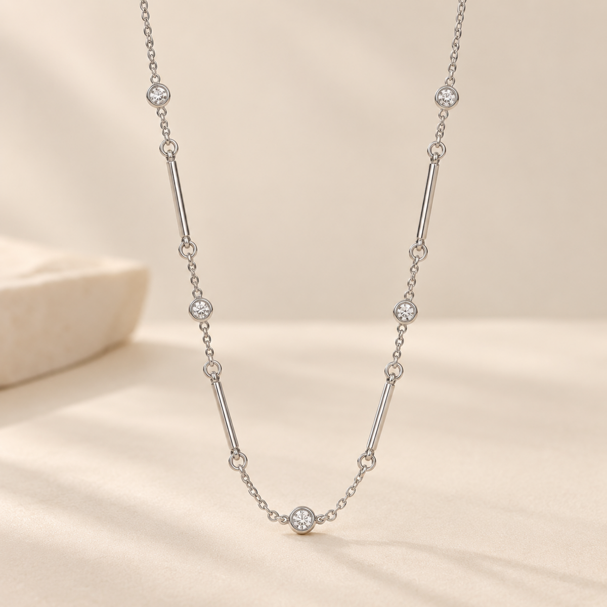 A delicate silver-toned chain necklace with alternating small round clear stones and cylindrical silver links, displayed on a soft cream fabric surface with a blurred stone prop in the background.