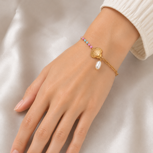 A gold-plated ladies bracelet featuring a gold paperclip chain on one side and a colorful beaded pearl strand with a shell charm on the other, shown on a model