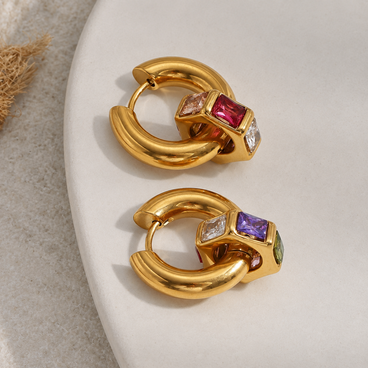 A pair of gold huggie hoop earrings with colorful gemstone accents, displayed on a white ceramic plate.