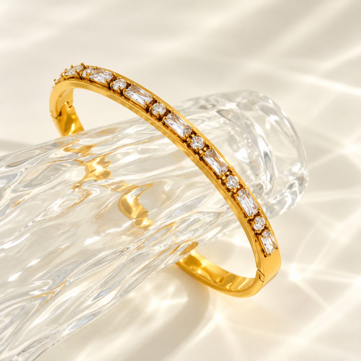 A gold hinged bangle bracelet with a row of clear baguette and round stones, displayed on a textured clear glass stand against a bright, sunlit cream background.