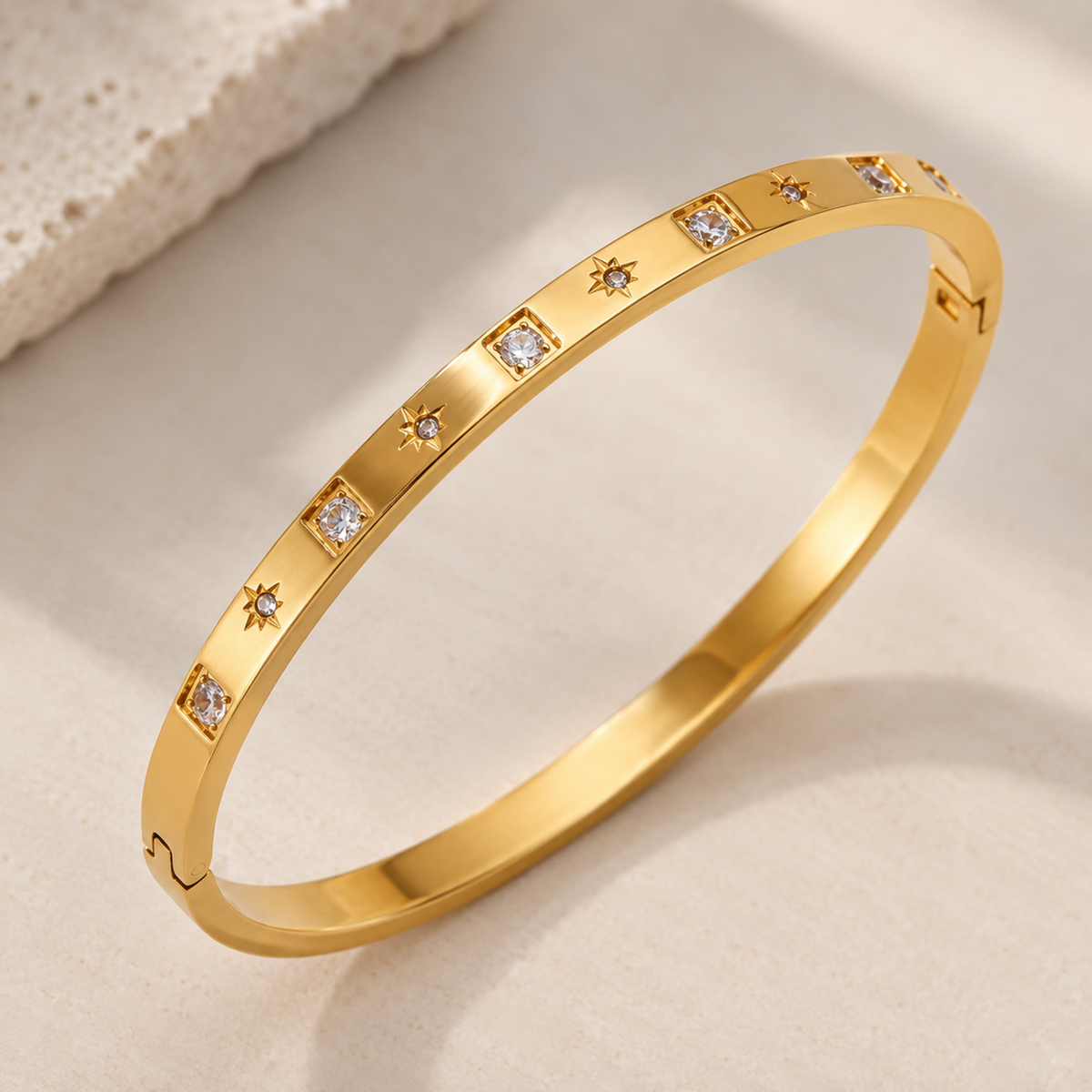 A polished gold bangle bracelet with small star-shaped diamond accents set along the outer band, photographed on a soft beige surface with a textured cream prop in the corner.