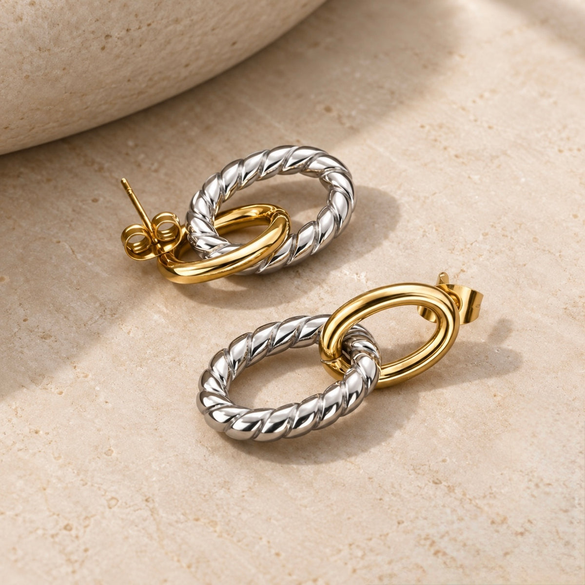 A pair of gold and silver drop earrings with twisted rope-style rings and smooth oval links, displayed on a light beige textured stone surface.