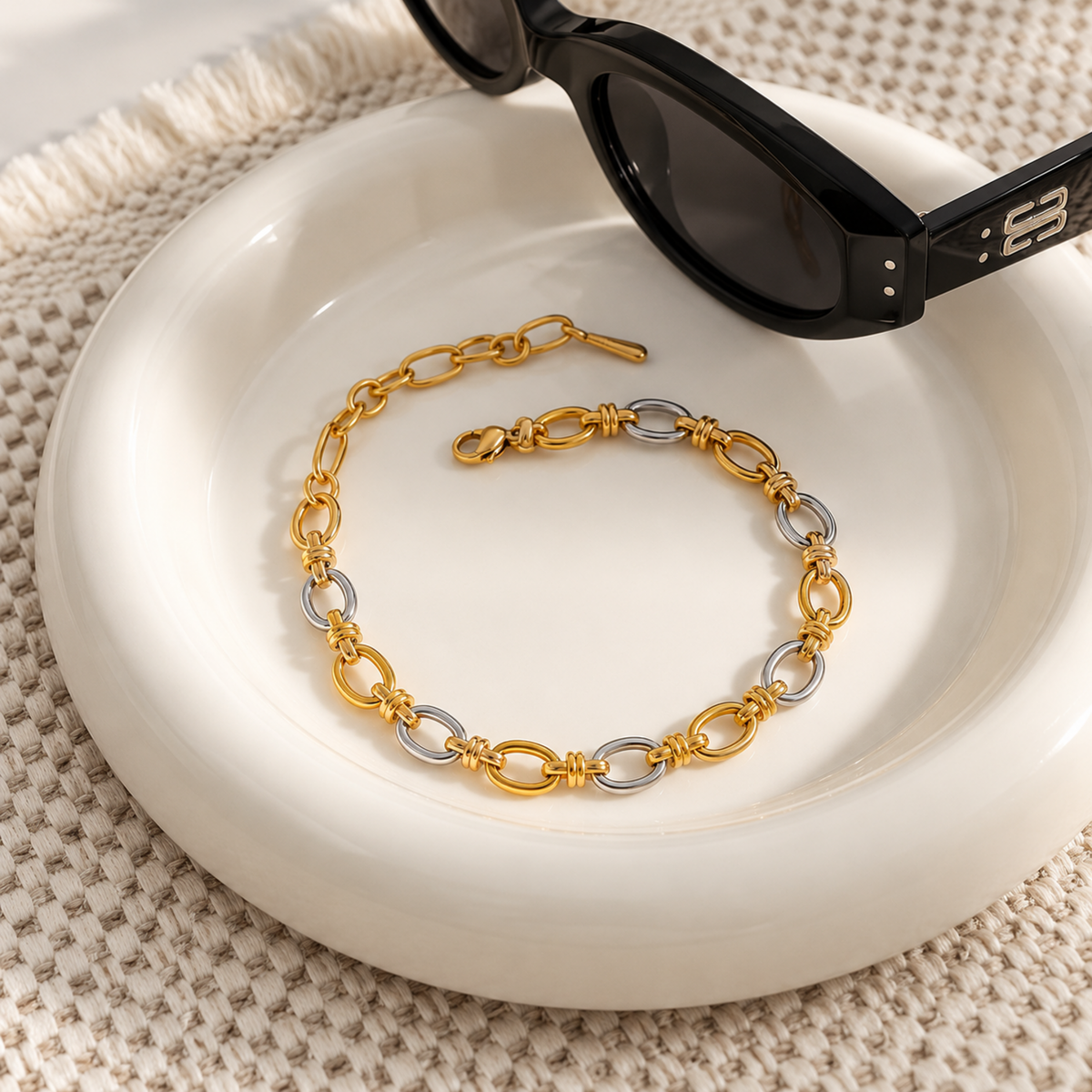 A gold and silver chain link bracelet is displayed on a white ceramic dish, with black sunglasses resting on the rim. The background is a textured beige woven mat.