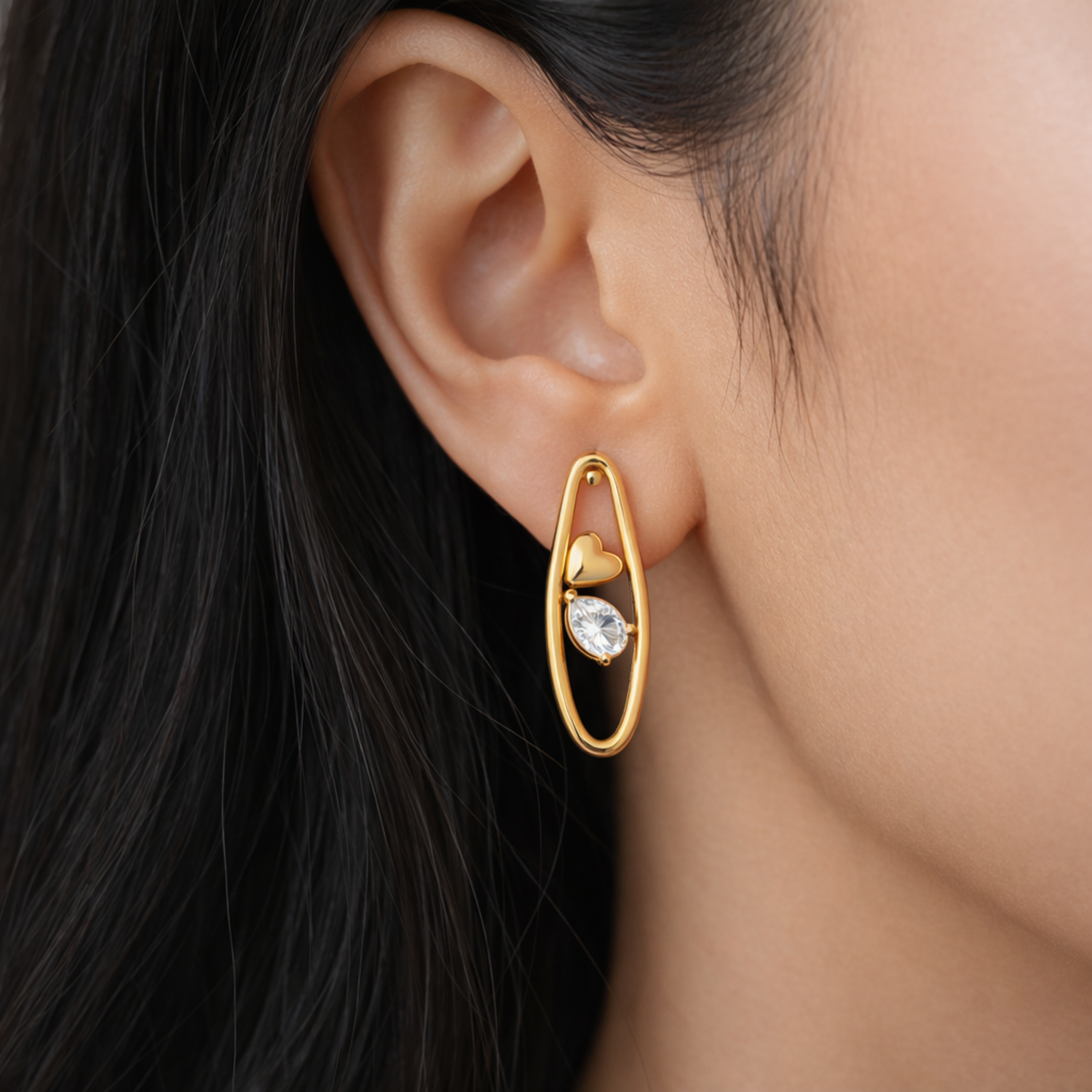 A gold-plated organic-shaped stud earrings for women featuring a small golden heart and a pear-cut crystal inside a golden loop, shown on a model