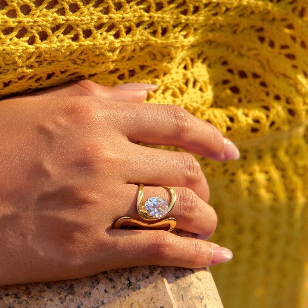 Close view of an artistic gold plated adjustable statement ring for women featuring a large round white stone set in a sculptural, wavy gold-tone band on a yellow textured background