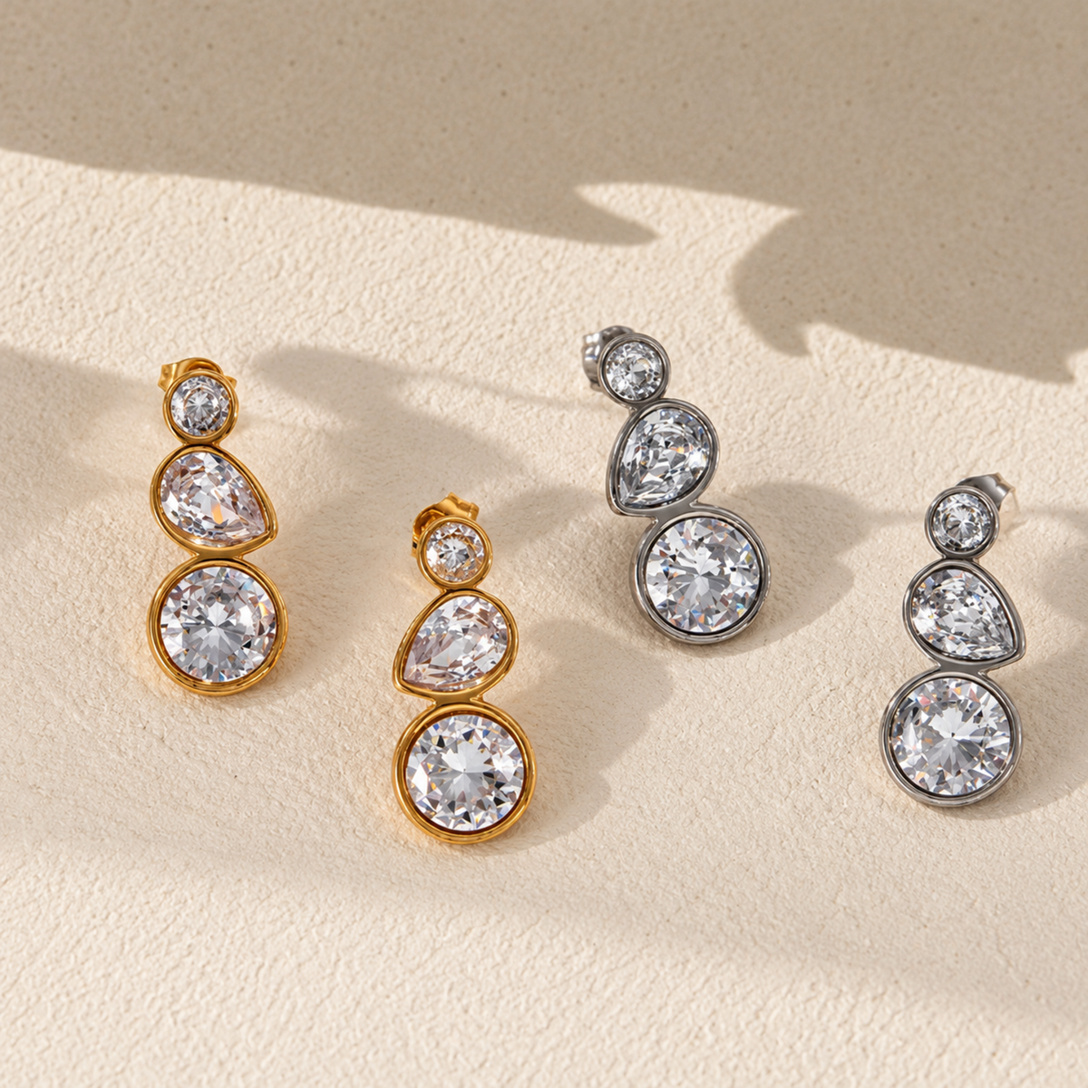 Four drop earrings with clear faceted gemstones in gold and silver-tone settings, arranged on a light beige textured surface with soft shadows.