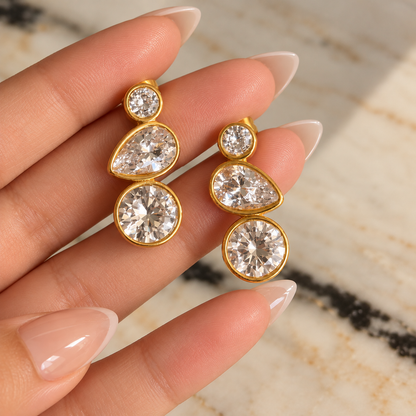 A close-up of a hand with manicured nails holding a pair of gold drop earrings with clear round and teardrop-cut gemstones, set against a light marble background.