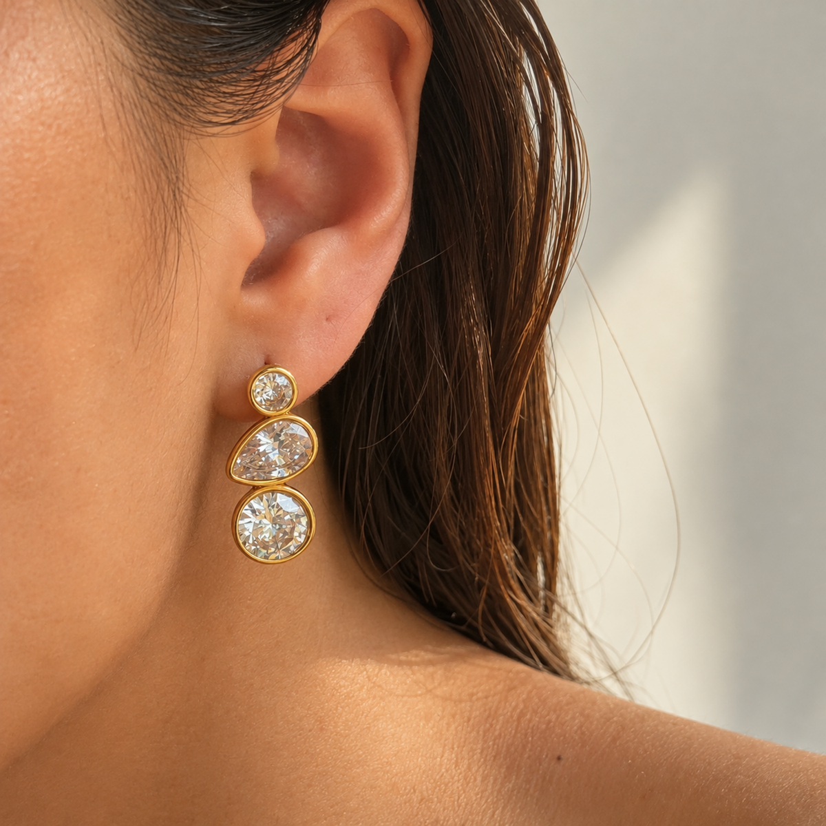A close-up of a model's ear wearing a gold drop earring with three clear round and teardrop gemstones.