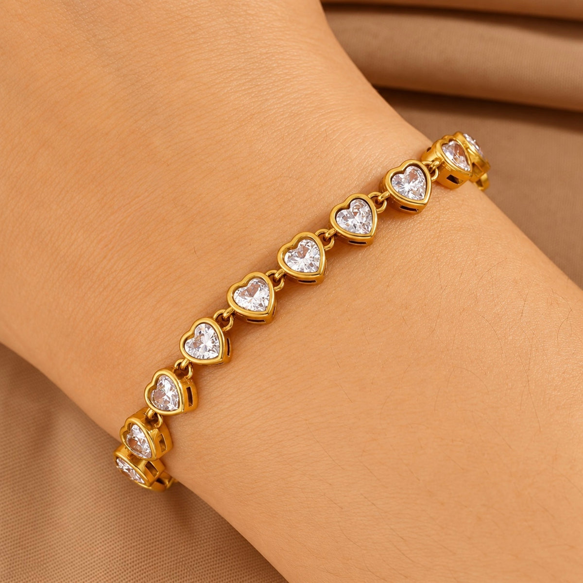 A gold heart-link bracelet with clear heart-shaped stones, shown worn on a wrist against a soft tan fabric background.