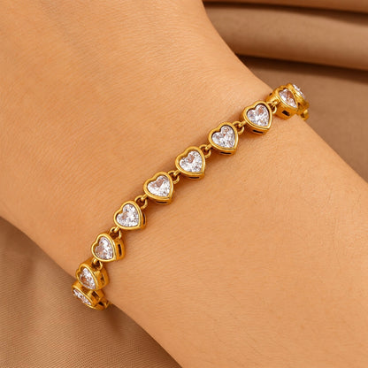 A gold heart-link bracelet with clear heart-shaped stones, shown worn on a wrist against a soft tan fabric background.
