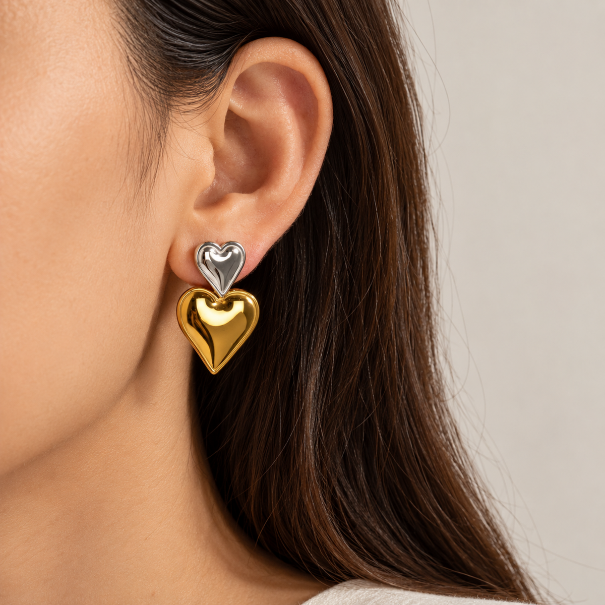 A close-up of a woman's ear wearing a double-heart stud earring with one shiny gold heart and one silver heart.