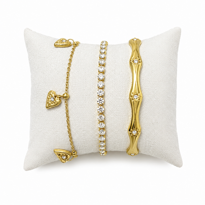 A three-piece gold-plated bracelet stack for women featuring an engraved wavy starburst bangle, a classic crystal tennis bracelet, and a fine link chain with three dangling gold heart charms