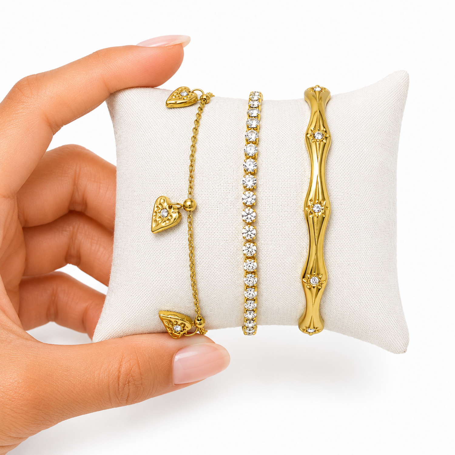 A close-view of a three-piece gold-plated bracelet stack for women featuring an engraved wavy starburst bangle, a classic crystal tennis bracelet, and a fine link chain with three dangling gold heart charms.