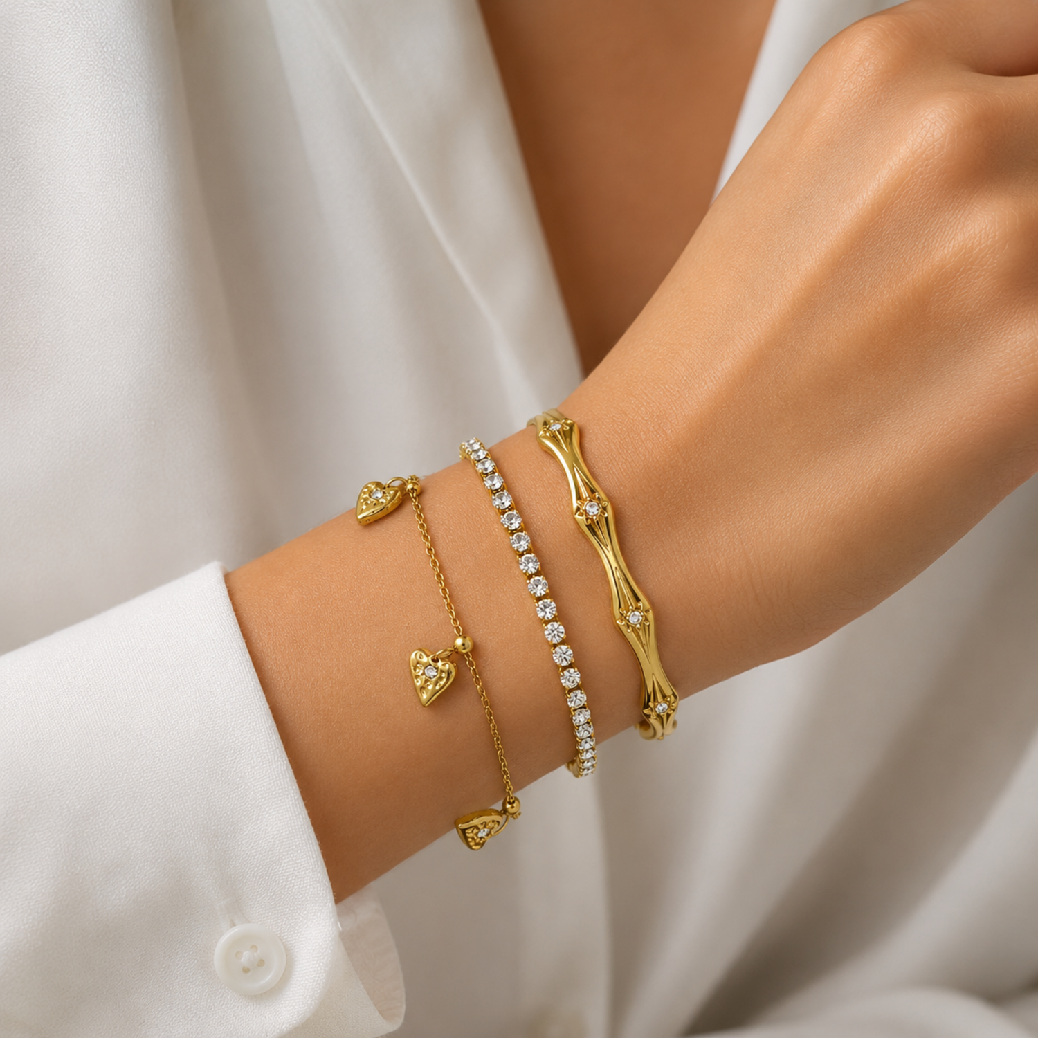 A three-piece gold-plated bracelet stack for women featuring an engraved wavy starburst bangle, a classic crystal tennis bracelet, and a fine link chain with three dangling gold heart charms, on a model