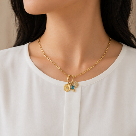A gold-plated stainless steel chain for women featuring paperclip links and a carabiner clasp with three hanging charms, including a turquoise stone and an evil eye, shown on a model