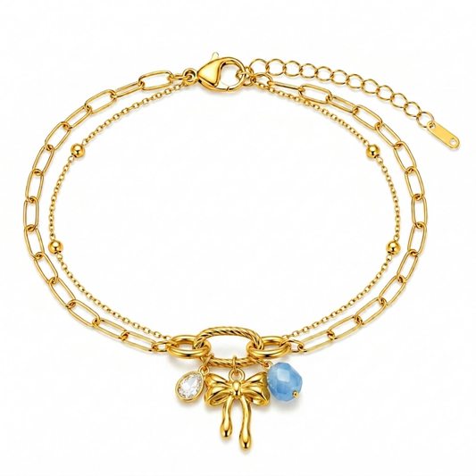 A gold-plated layered chain bracelet for women featuring a paperclip chain, a satellite chain, a gold bow charm, and a blue stone