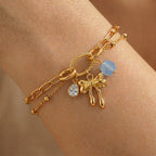 A gold-plated layered chain bracelet for women featuring a paperclip chain, a satellite chain, a gold bow charm, and a blue stone, shown on a model