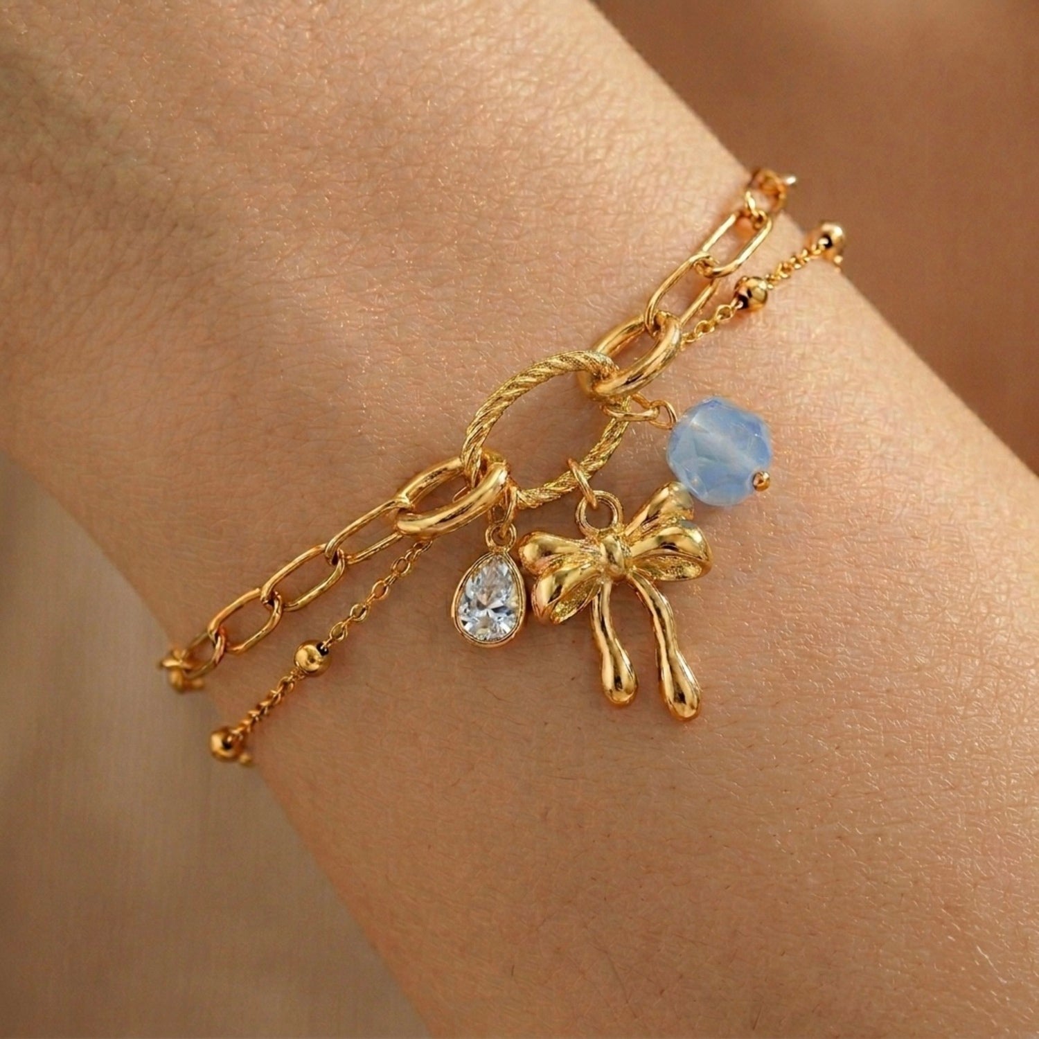 A gold-plated layered chain bracelet for women featuring a paperclip chain, a satellite chain, a gold bow charm, and a blue stone, shown on a model
