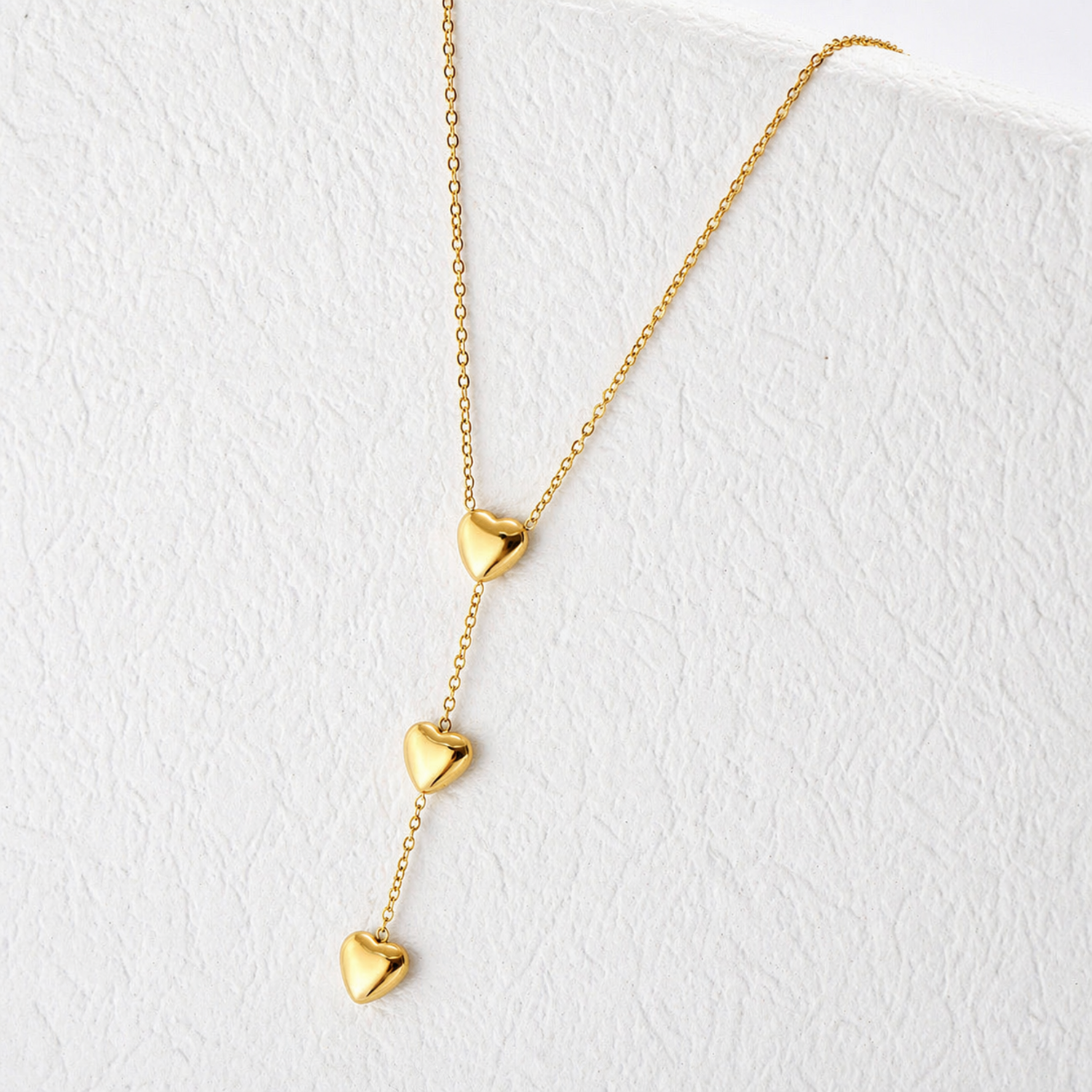 A gold-plated stainless steel chain for women featuring a cascading vertical drop of three polished heart pendants, shown on a textured white surface