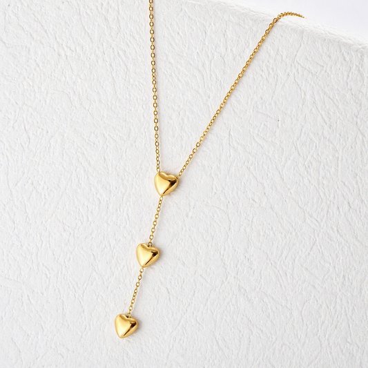 A gold-plated stainless steel chain for women featuring a cascading vertical drop of three polished heart pendants, shown on a textured white surface