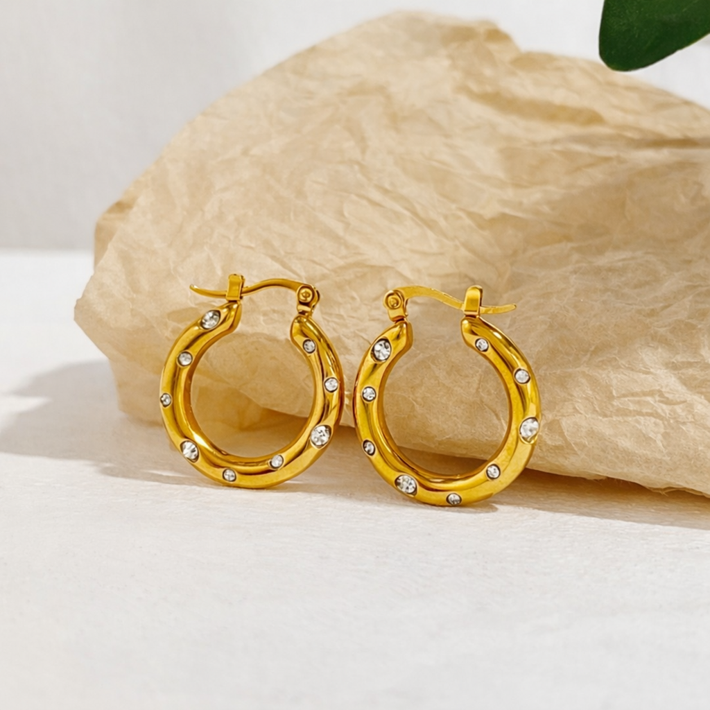 A pair of gold-plated thick hoop earrings for women featuring scattered clear crystals, shown against a textured surface