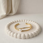Modern gold-plated double-band adjustable cuff bracelet for women featuring a single central pearl, resting on a white bobbled jewelry dish.
