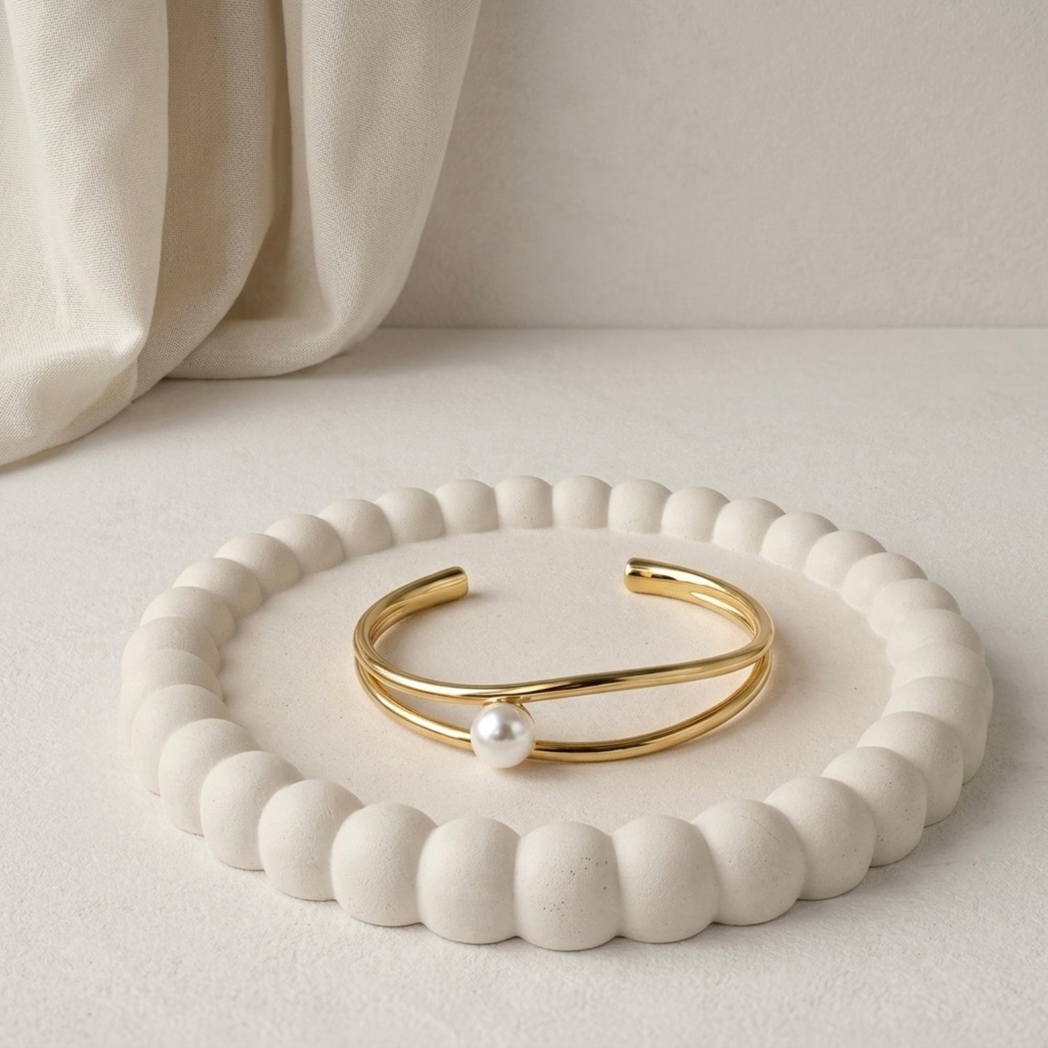Modern gold-plated double-band adjustable cuff bracelet for women featuring a single central pearl, resting on a white bobbled jewelry dish.