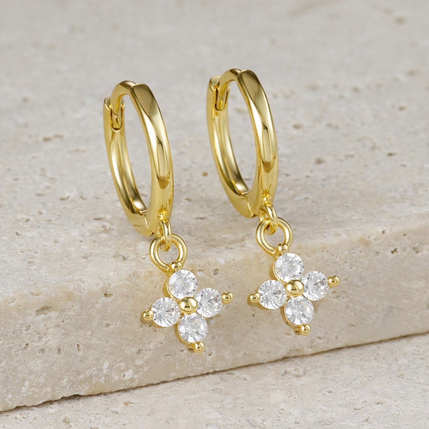 A pair of gold-plated hoop earrings for women featuring a sparkling four-petal crystal clover drop, shown on a textured surface