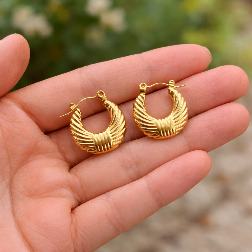 A pair of gold-plated textured croissant-style hoop earrings for women with a ribbed dome design, shown held in a hand.