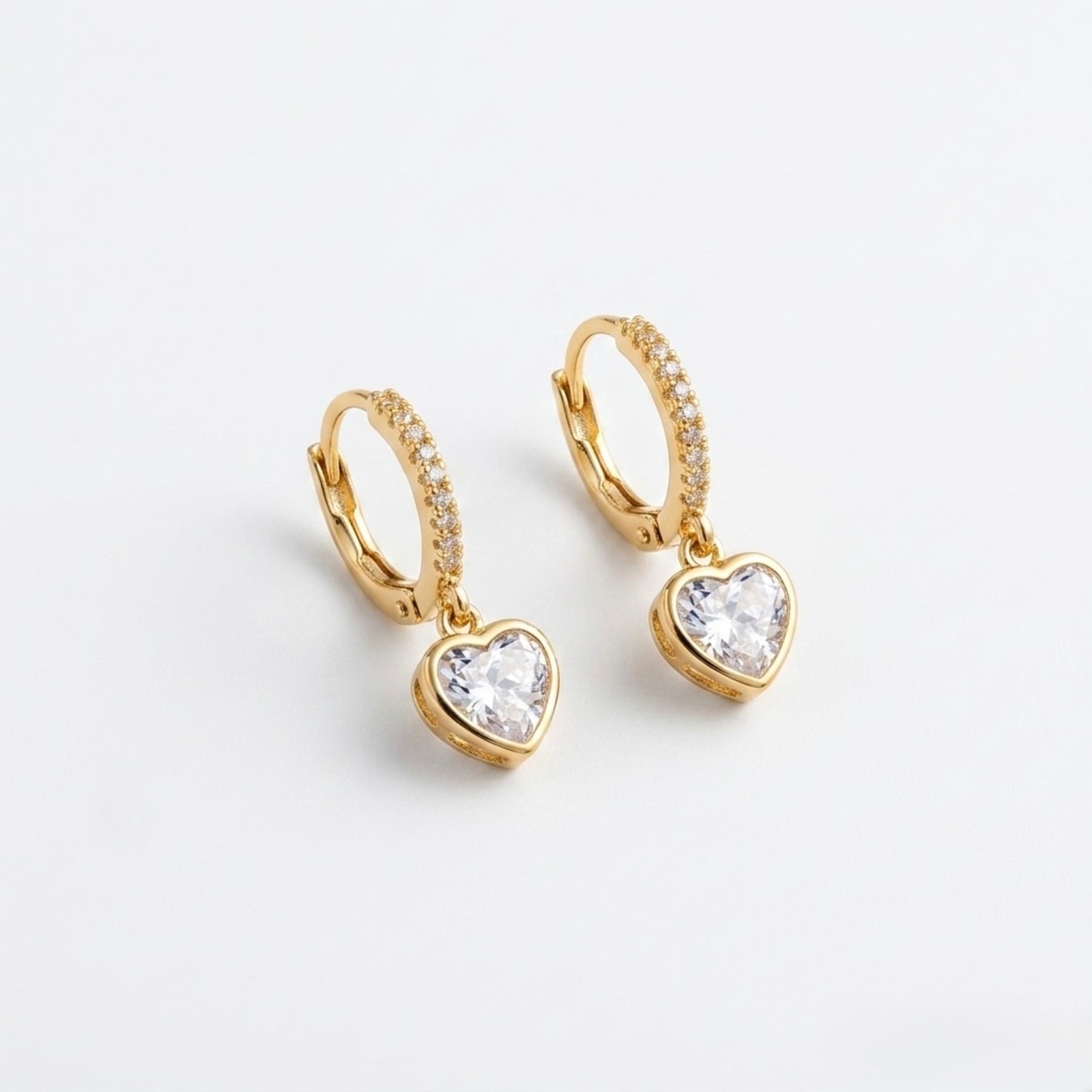 A close-view of a pair of gold-plated hoop earrings for women featuring a suspended heart-shaped clear crystal drop, shown on a clean white background.
