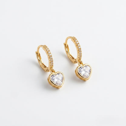 A close-view of a pair of gold-plated hoop earrings for women featuring a suspended heart-shaped clear crystal drop, shown on a clean white background.