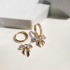 A pair of gold-plated hoop earrings for women featuring a crystal bow drop with heart-shaped stones, shown on a white background.