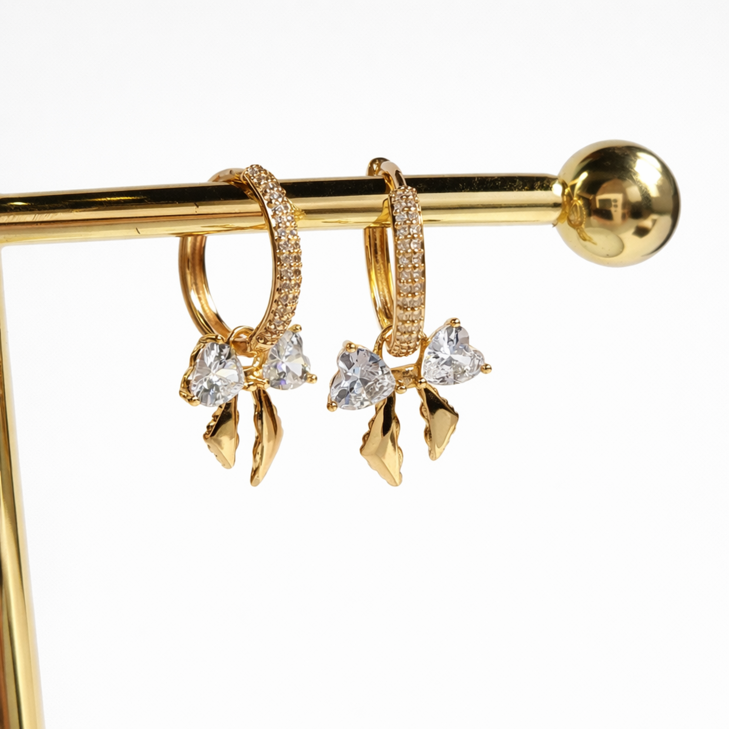 A close-view of a pair of gold-plated hoop earrings for women featuring a crystal bow drop with heart-shaped stones, shown on a white background