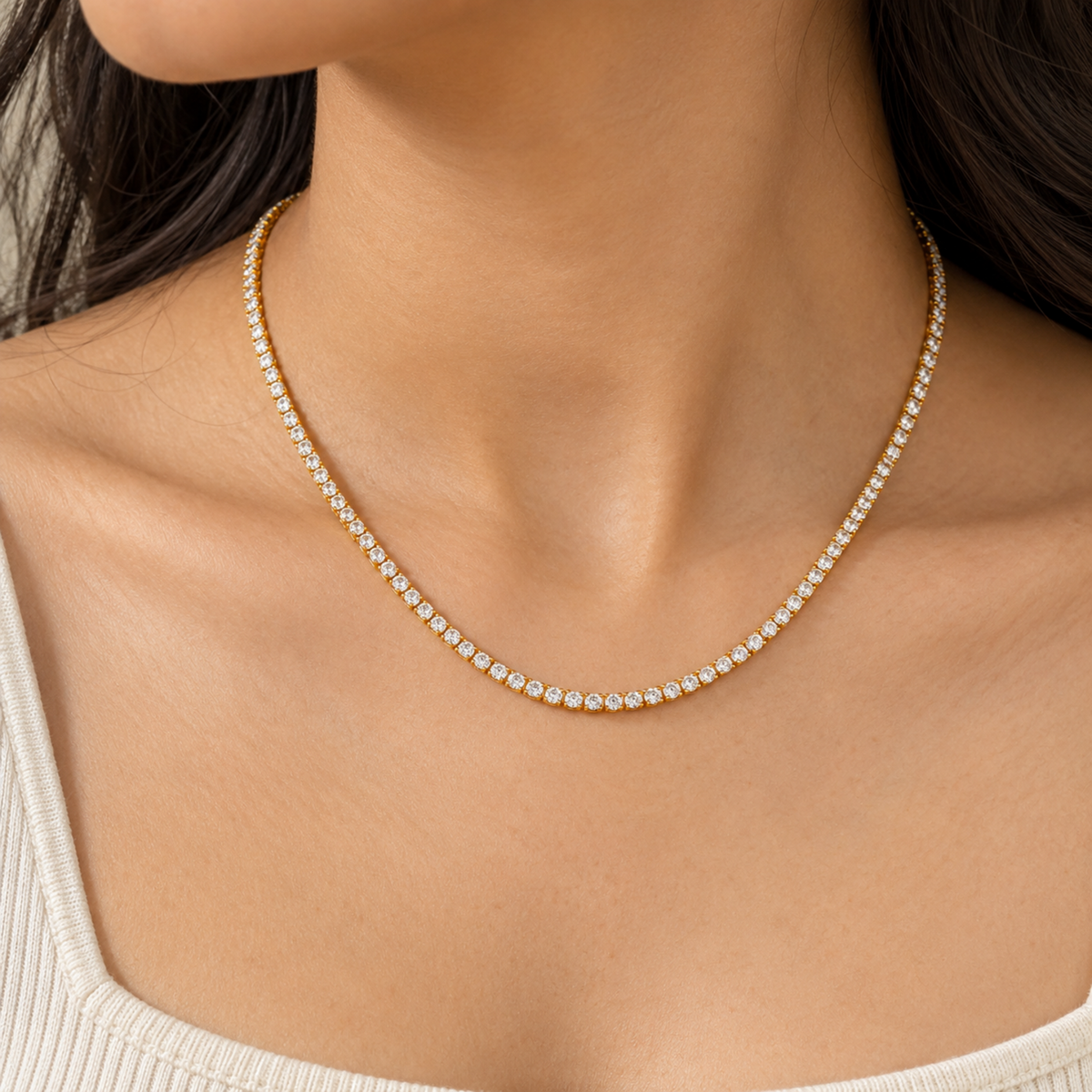 A close-up of a woman's neck and upper chest wearing a delicate gold tennis necklace with small clear stones, paired with a light beige ribbed top.