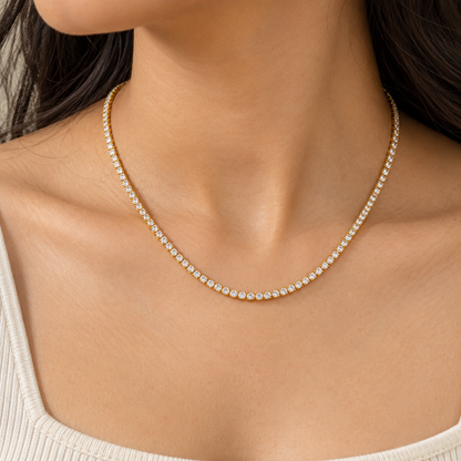 A close-up of a woman's neck and upper chest wearing a delicate gold tennis necklace with small clear stones, paired with a light beige ribbed top.