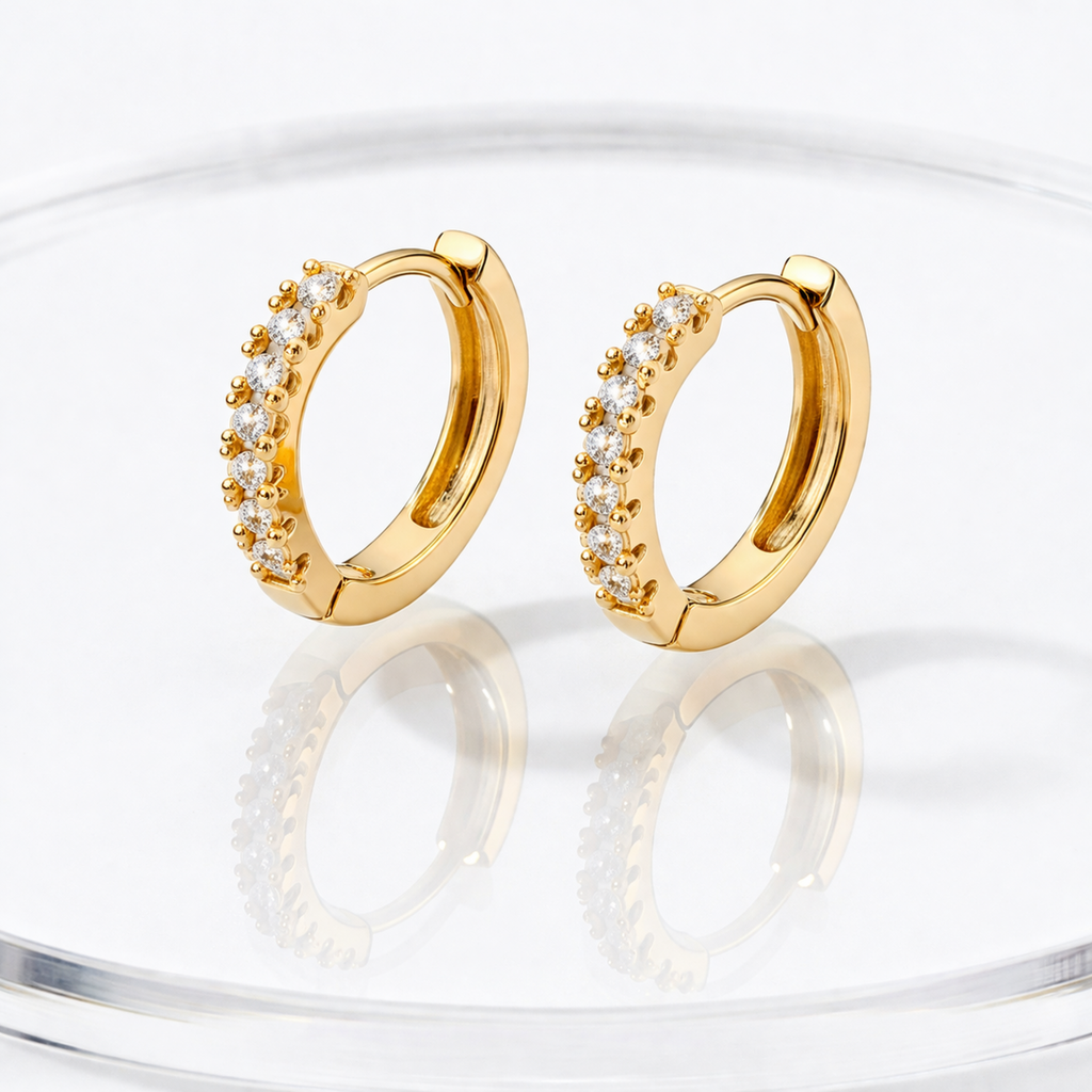A pair of gold-plated huggie hoop earrings for women featuring a delicate row of clear crystals, shown reflecting on a glass surface.
