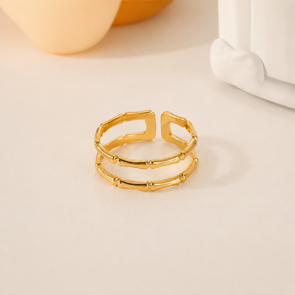 A gold-plated open ring with a textured, segmented bamboo-like design, displayed on a light beige surface with soft-focus background elements.