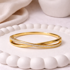 A luxurious gold-plated double-band cross-over bangle for women featuring one polished strand and one strand of brilliant pavé white stones displayed on a ceramic tray