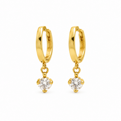 A pair of gold huggie hoop earrings with a dangling heart-shaped clear gemstone, shown on a plain white background.