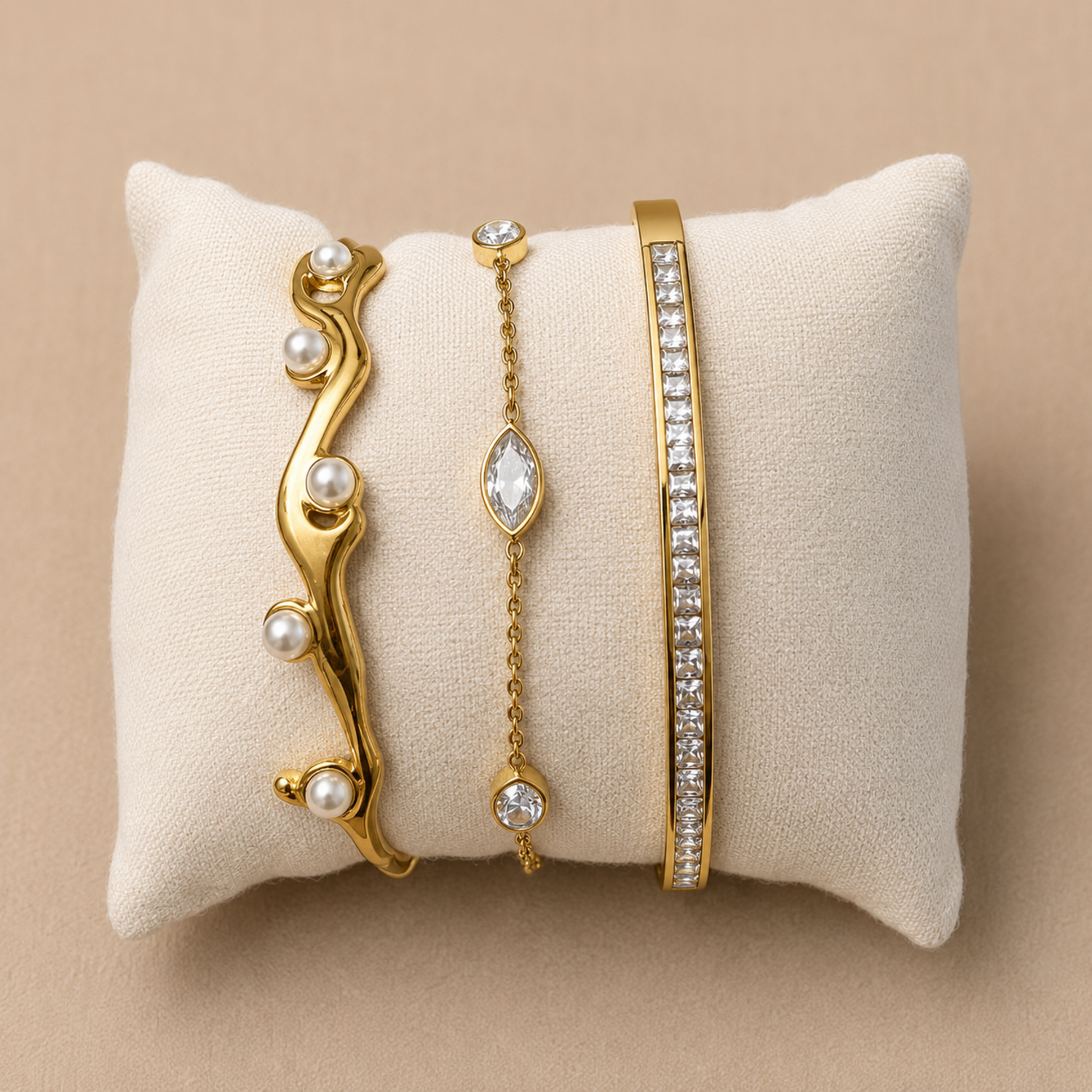 Three gold bracelets with pearls and crystals on a beige pillow against a beige background