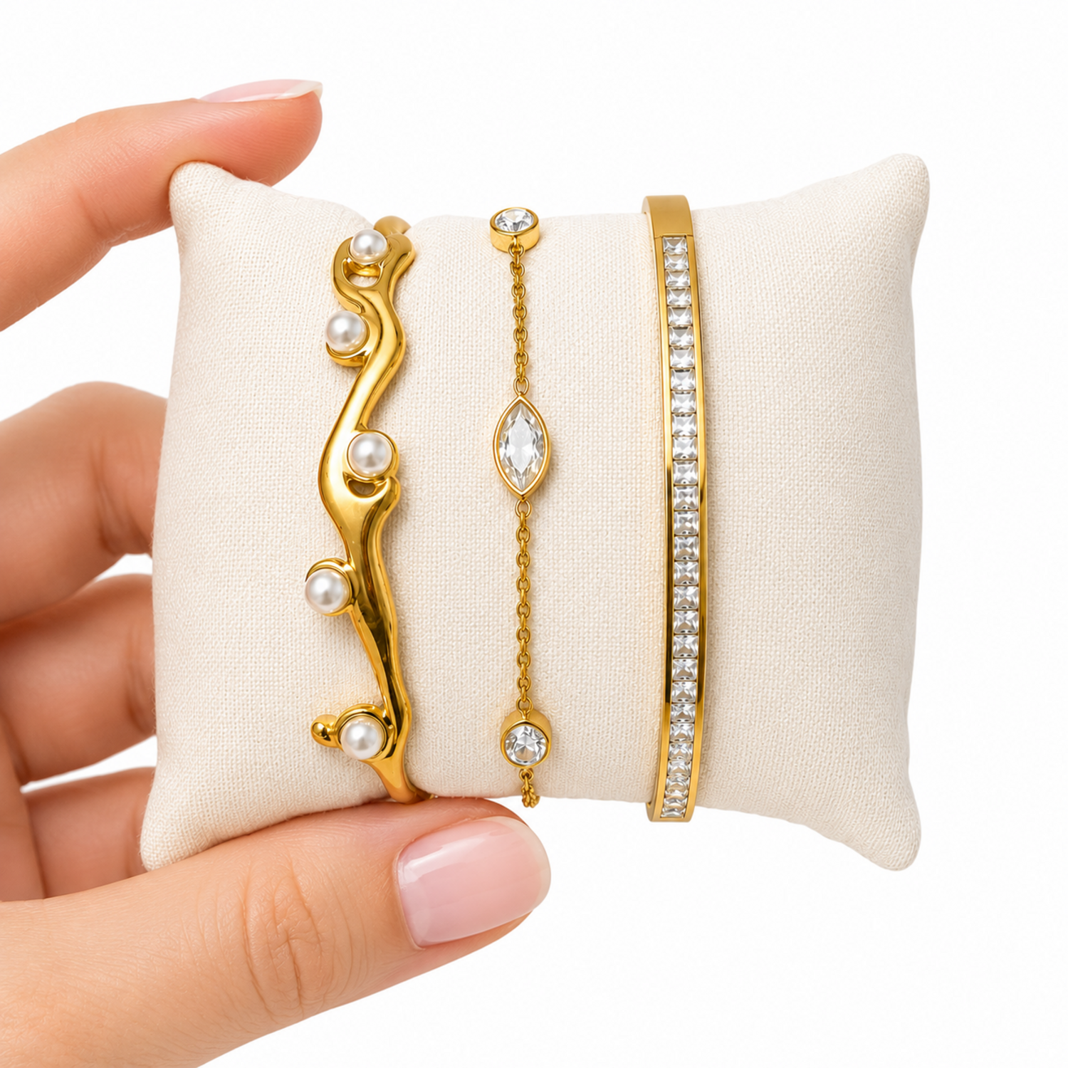 Three gold bracelets on a white cushion held by a hand with pink nail polish.
