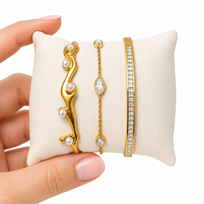 Three gold bracelets on a white cushion held by a hand with pink nail polish.