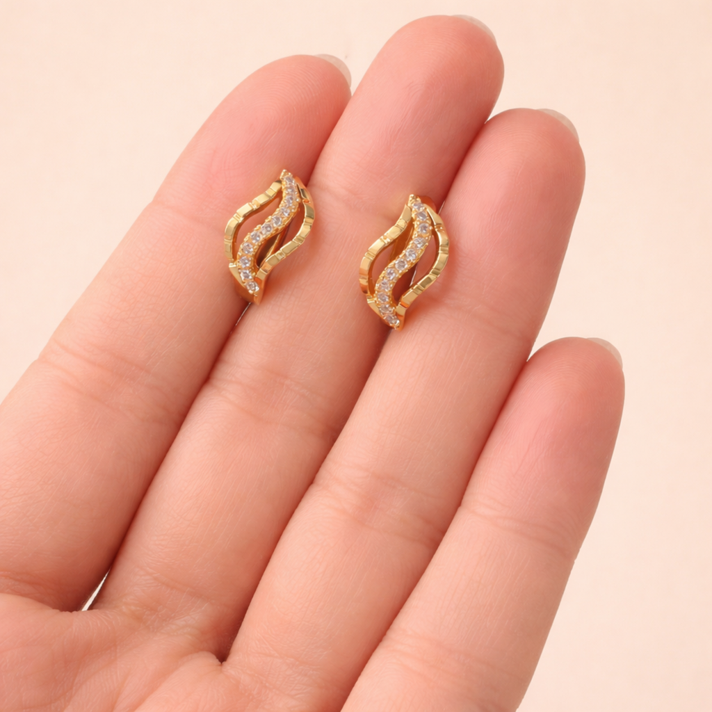 A pair of gold-plated wave-shaped huggie hoop earrings for women featuring a curved row of clear crystals, shown on a hand