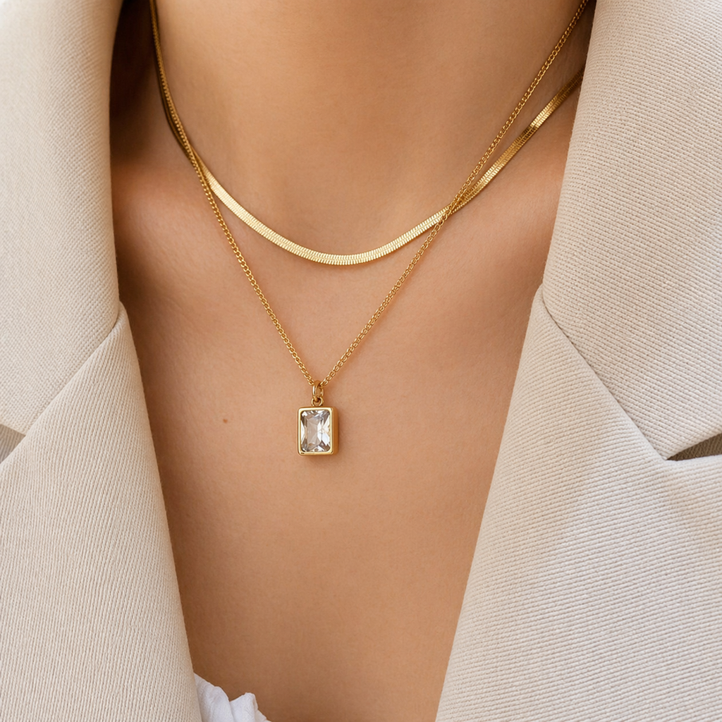 A gold-plated stainless steel chain for women featuring a layered necklace design with a flat herringbone chain and a link chain holding an emerald-cut crystal pendant