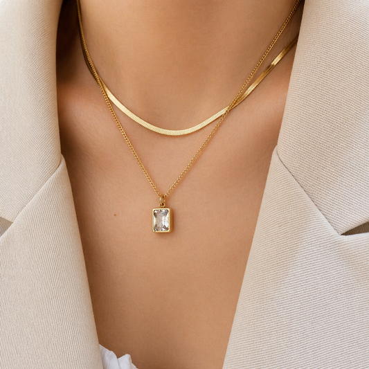 A gold-plated stainless steel chain for women featuring a layered necklace design with a flat herringbone chain and a link chain holding an emerald-cut crystal pendant
