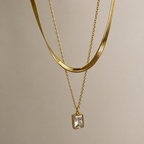 A close-view of a gold-plated stainless steel chain for women featuring a layered necklace design with a flat herringbone chain and a link chain holding an emerald-cut crystal pendant