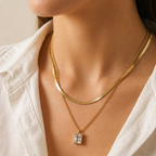 A gold-plated stainless steel chain for women featuring a layered necklace design with a flat herringbone chain and a link chain holding an emerald-cut crystal pendant shown on a model