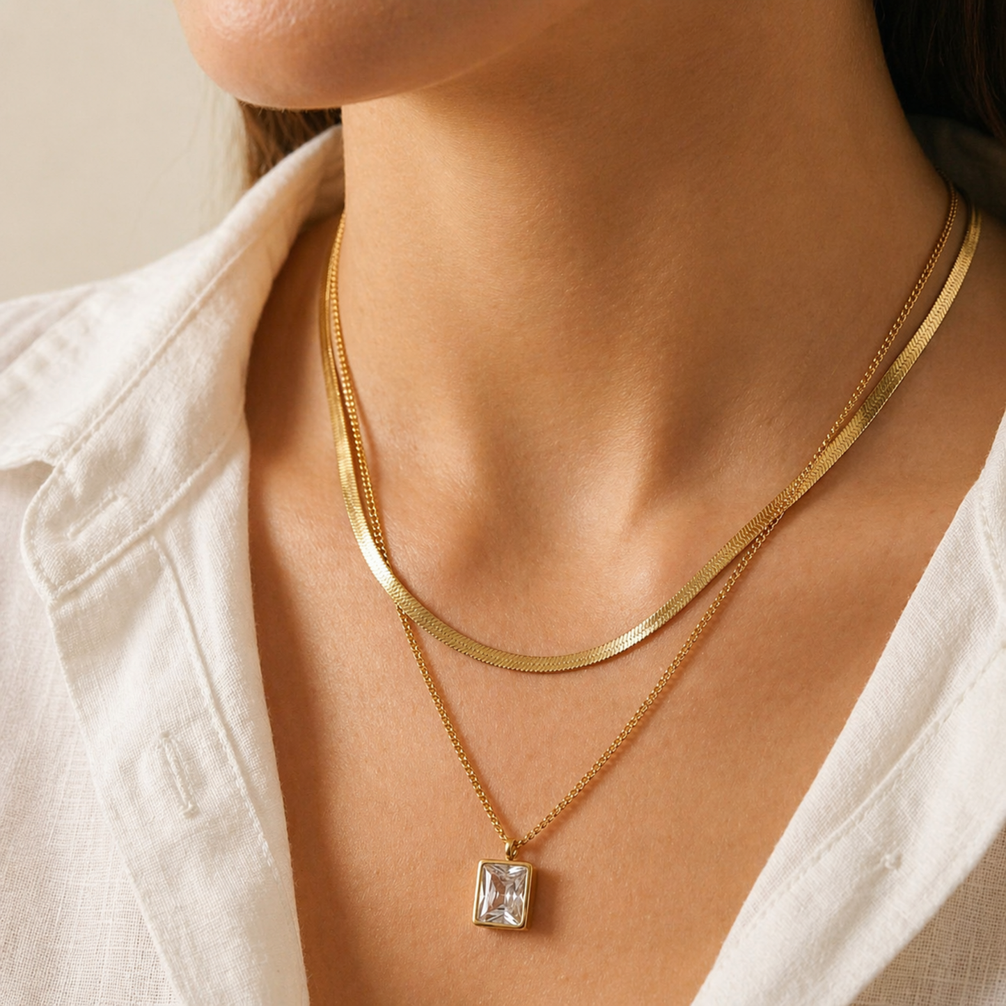 A gold-plated stainless steel chain for women featuring a layered necklace design with a flat herringbone chain and a link chain holding an emerald-cut crystal pendant shown on a model