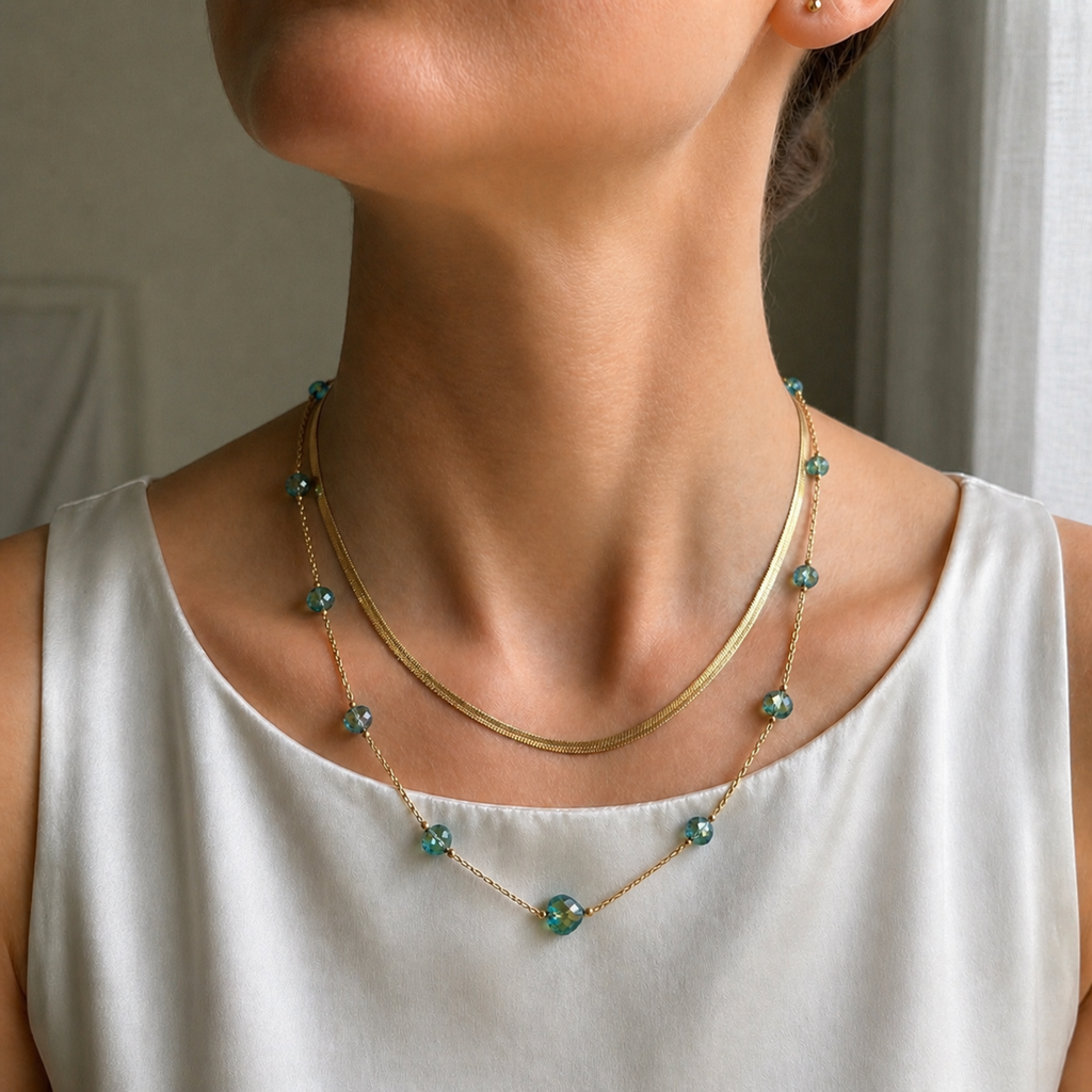 A gold-plated stainless steel chain layered necklace for women featuring a smooth snake chain and a second chain with green faceted heart crystals, shown on a model